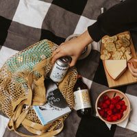 Person sitting on picnic blanket with raspberries, cheese and crackers. Hand reaching for Stoble cold brew can that is in a yellow knit totebag with a book.. 