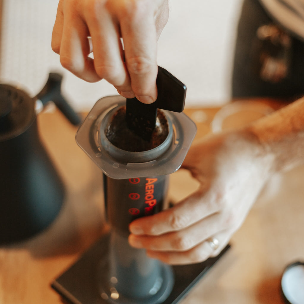 Hand stabilizing Aeropress coffee maker while other hand holds stirring stick and stirs the coffee in Aeropress.